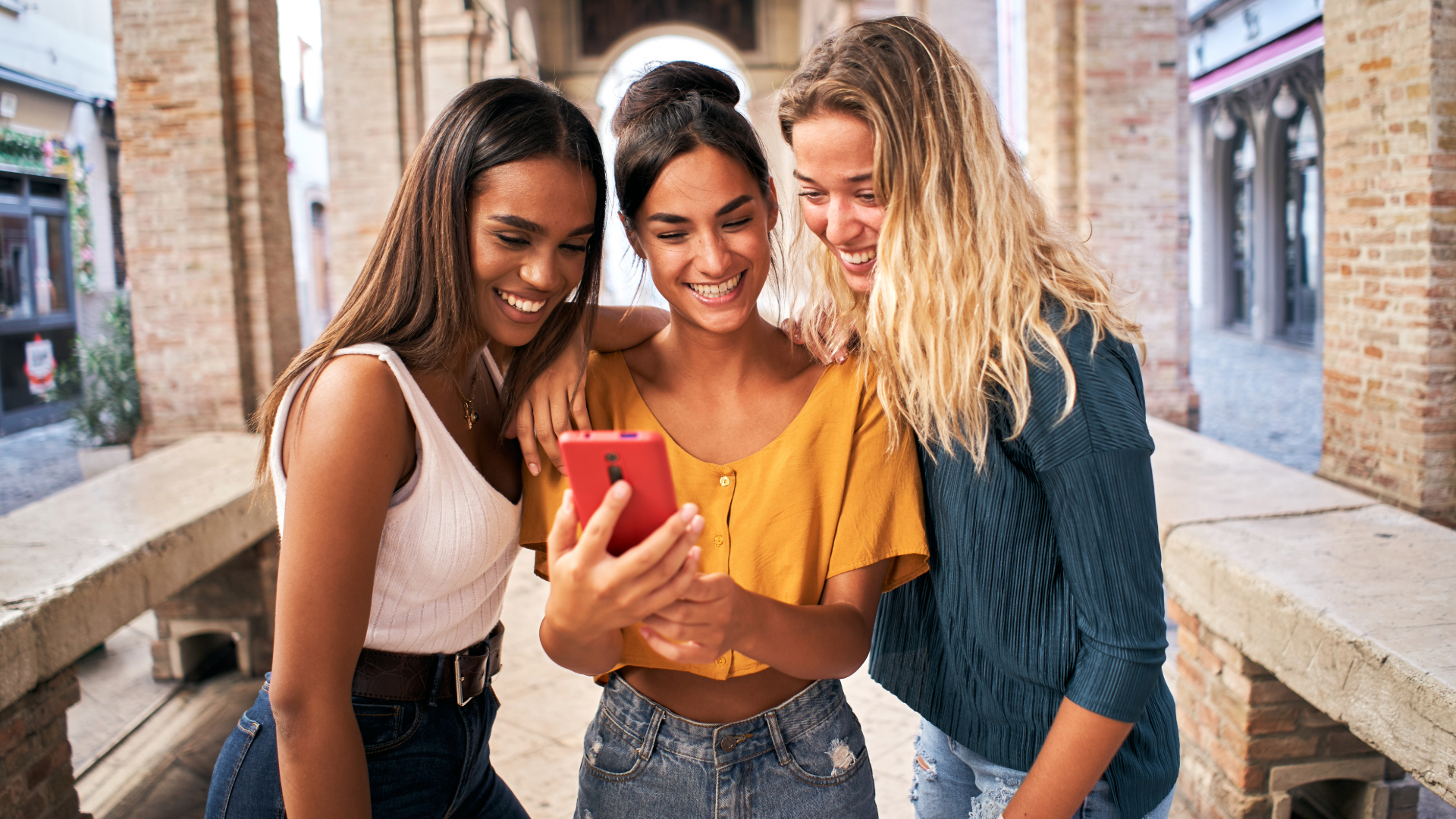 Three young women gathered together, exploring user profiles on wrthy app at a cell phone.